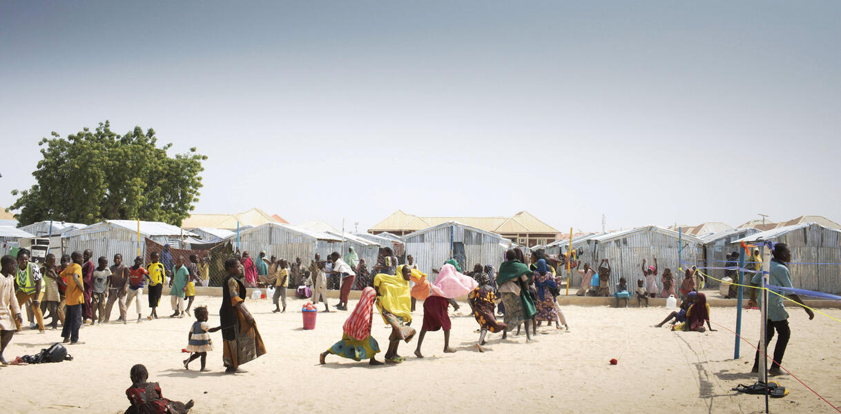 Sandiger Wüstenboden in Nigeria, auf dem viele Menschen sind, Erwachsene und Kinder, manche laufen, manche sitzen im Sand, im Hintergrund die Zelte eines Flüchtlingslagers.