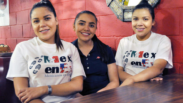 Die drei Schwestern Citlali, Deisy und Paola Alvarado sitzen lächelnd  nebeneinander an einem Tisch, zwei von ihnen sind eineiige Zwillinge