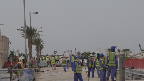 Mehrere Männer in Arbeitskleidung stehen auf einer Baustelle mit Palmen im Hintergrund