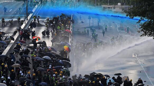 Demonstrierende Menschen, die sich mit Regenschirmen vor Angriffen der Polizei mit Wasserkanonen schützen. 