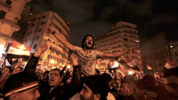Das Bild zeigt die Szene einer Demonstration bei Nacht: Viele Menschen schwenken ägyptische Flaggen, ein Mann sitzt auf den Schultern eines anderen und hebt beide Hände in die Höhe