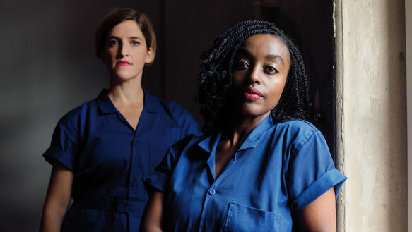 Eine weiße und eine schwarze Frau tragen blaue Overalls oder Arbeitshemden und stehen im Halbdunkel eines Raumes in den Licht fällt.