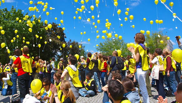 An einem sonnigen Tagen lässt eine große Menge Menschen, die Westen mit dem Amnesty-Logo tragen, Luftballons in den Himmel steigen.