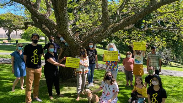 Sechzehn Personen mit Masken stehen und sitzen in einem Park unter einem Baum und halten teilweise gelbe Schilder vor sich.