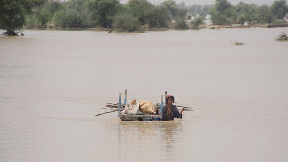 Eine Frau watet mit ein paar Habseligkeiten durch eine überschwemmte Landschaft mit hüfthohem Wasser.