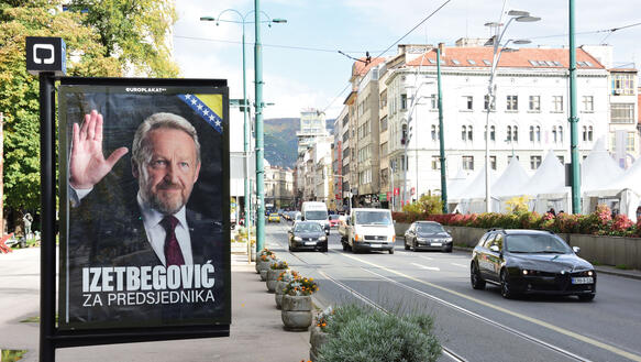 Eine Straße, auf der ein Wahlplakat aufgehängt ist, das für einen politischen Kandidaten wirbt, "Izetbegovic za Predsjednika" steht darauf, über die Straße mit Straßenbahnschienen fahren Autos.