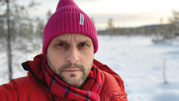 Ein Mann mit Dreitagebrat trägt Mütze und Schal in einer verschneiten Landschaft.