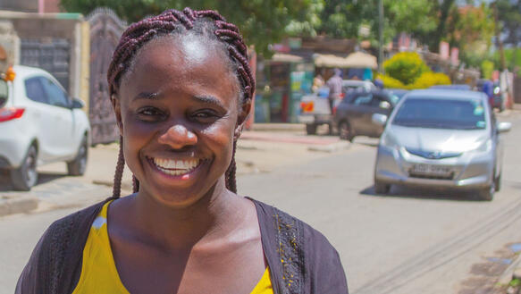 Eine afrikanische Frau steht au einer Straße und lächelt, die Sonne scheint, hinter ihr fahren und parken Autos.