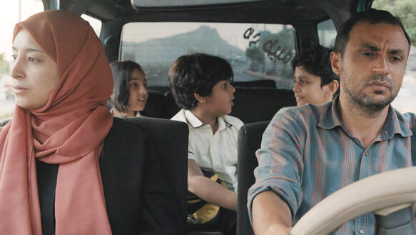 Eine Familie sitzt in einem Auto, der Mann fährt, die Frau auf dem Beifahrersitz, sie trägt ein Kopftuch, auf der Rückbank sitzen drei Kinder.