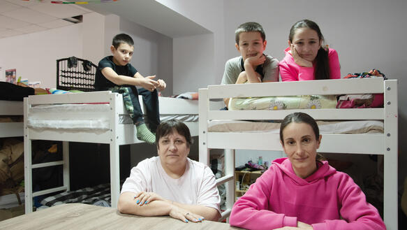 Eine Familie in einem Zimmer einer Flüchtlingsunterkunft, zwei Hochbetten, darauf liegend und sitzend drei Kinder, ein Junge und zwei Mädchen, vor den Hochbetten am Tisch zwei Frauen, sitzend.