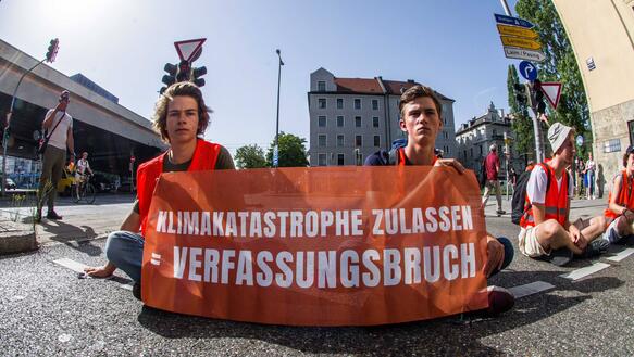 Das Bild zeigt zwei junge Männer, die auf einer Straßenkreuzung sitzen und ein Transparent mit der Aufschrift "Klimakatastrophe zulassen ist gleich Verfassungsbruch".