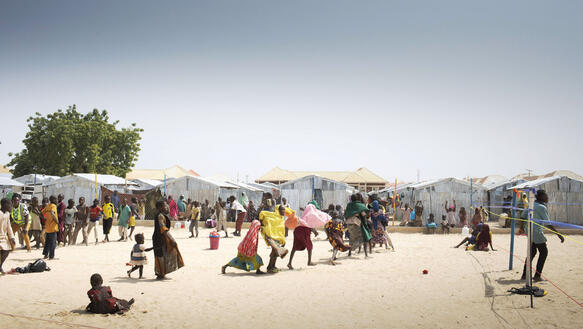 Sandiger Wüstenboden in Nigeria, auf dem viele Menschen sind, Erwachsene und Kinder, manche laufen, manche sitzen im Sand, im Hintergrund die Zelte eines Flüchtlingslagers.